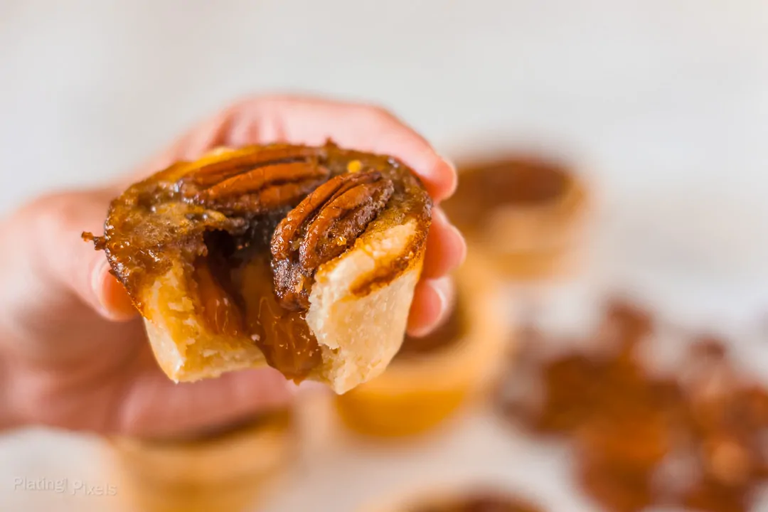 Hand holding a pecan tassie with a bite taken out of it and caramel filling oozing out