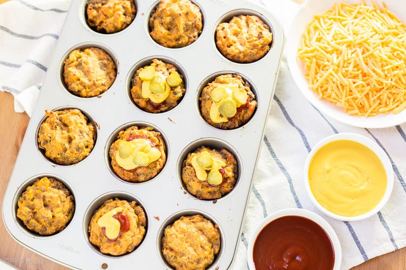 Mini Cheeseburger Muffins in a muffin pan with grated cheese, ketchup and mustard at the side