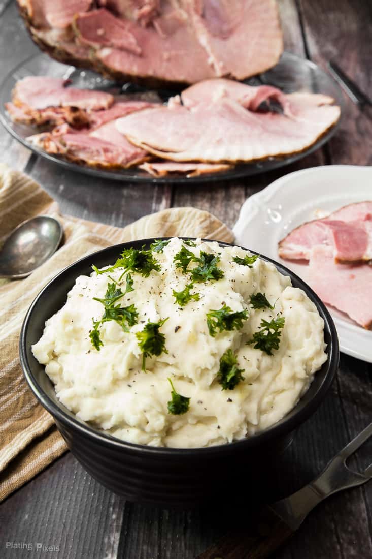 Creamy Mashed Potatoes in a black bowl garnished with green herbs on a table with carved baked ham