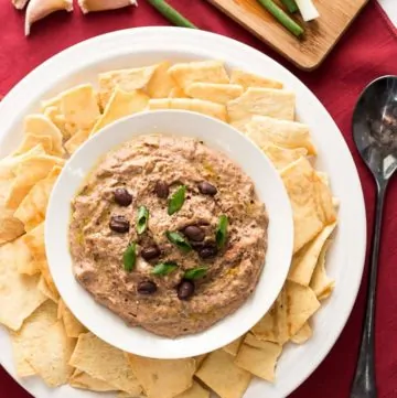 Sun Dried Tomato Black Bean Hummus in a bowl with chips around it and garlic cloves at the side