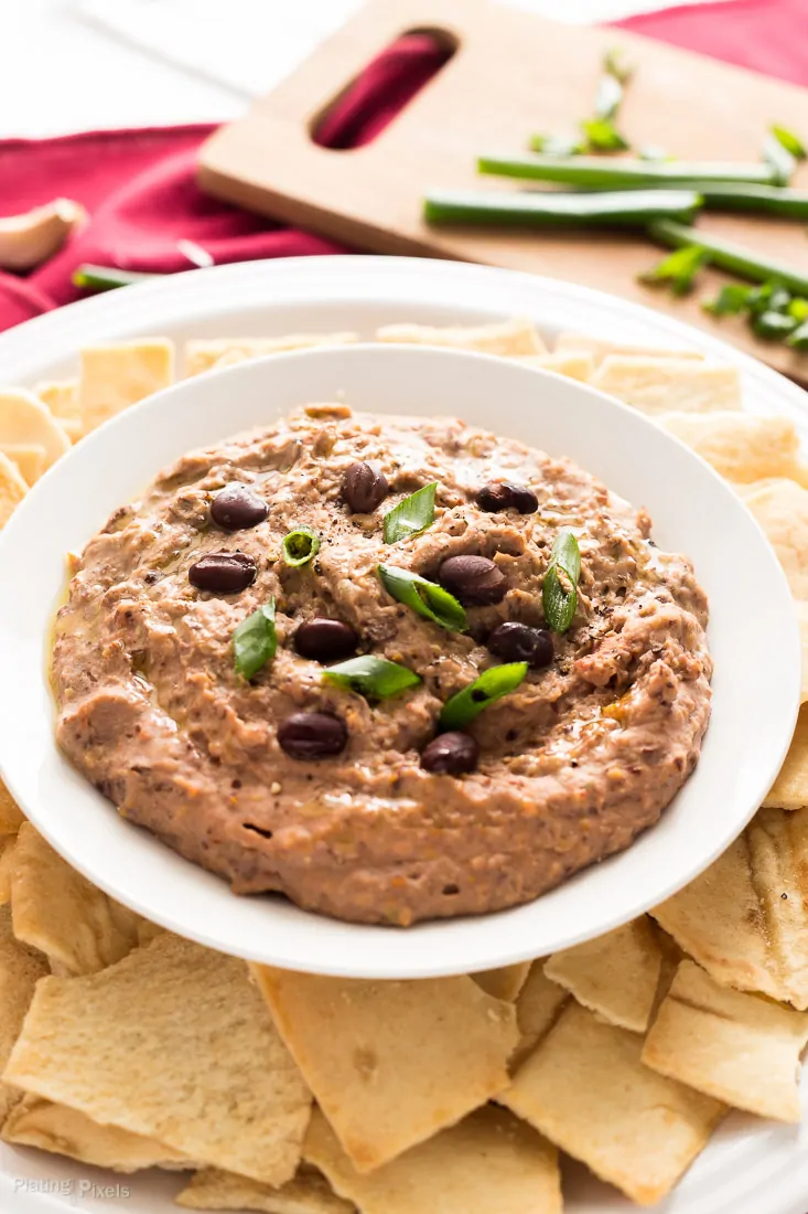 A close up of Sun Dried Tomato Black Bean Hummus in a bowl with chips around it and garnished with green onions