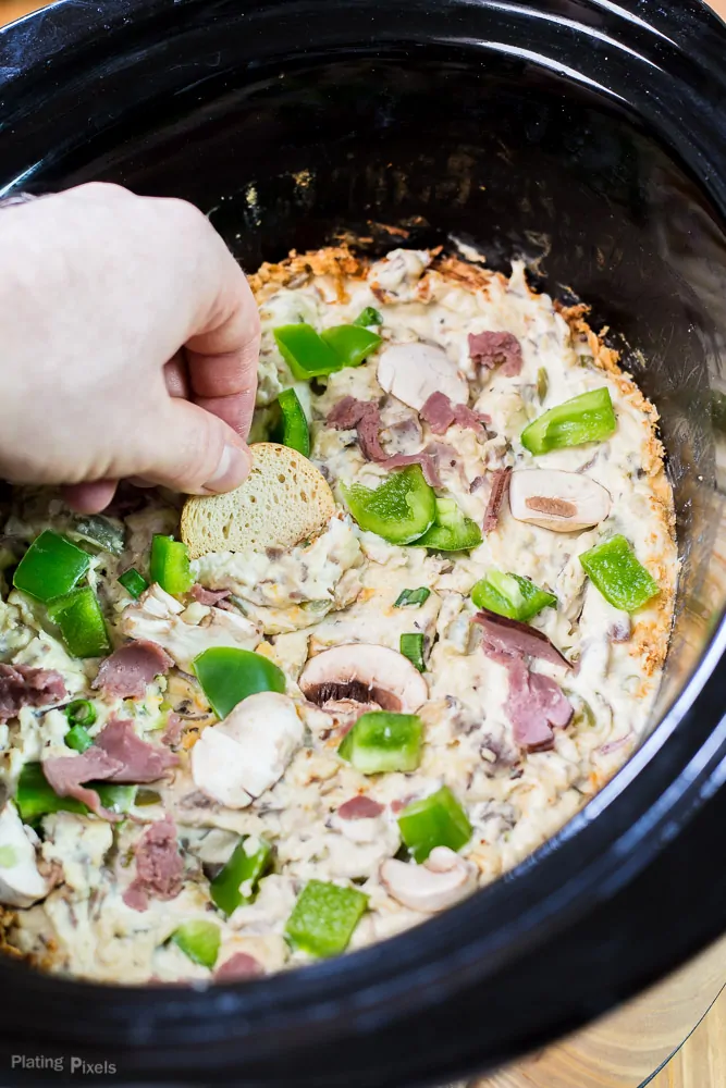 A hand dunking a cracker into a Slow Cooker Philly Cheese Steak dip