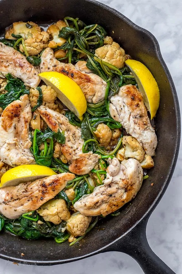 Process shot of lemon chicken cooking in a skillet with wedges of lemon and spinach