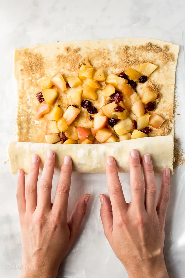 Rolling a puff pastry sheet topped with brown sugar and cooked apple pieces