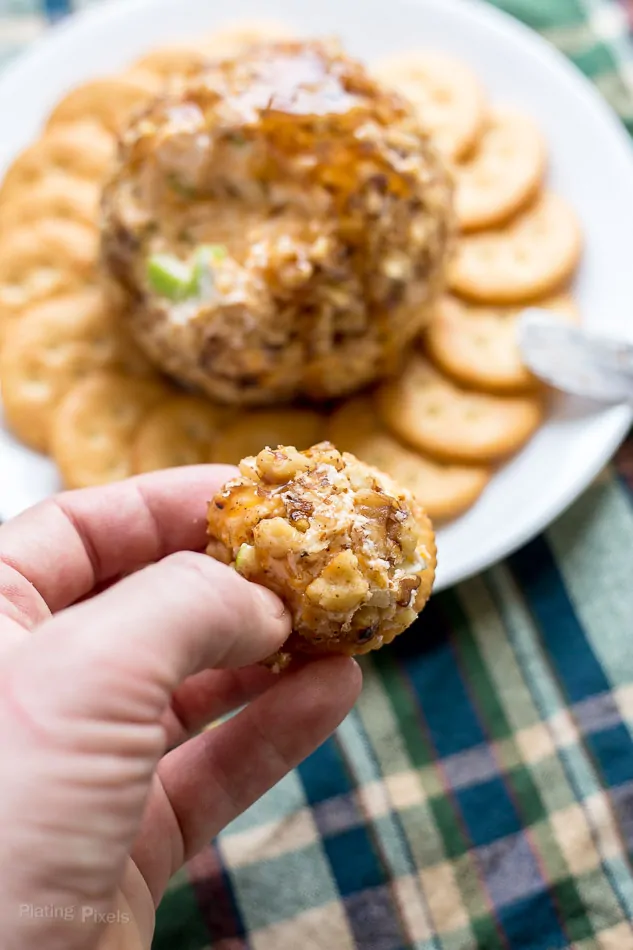 A hand holding some cheese ball spread on a cracker and a Caramel Apple Cheese Ball in the background