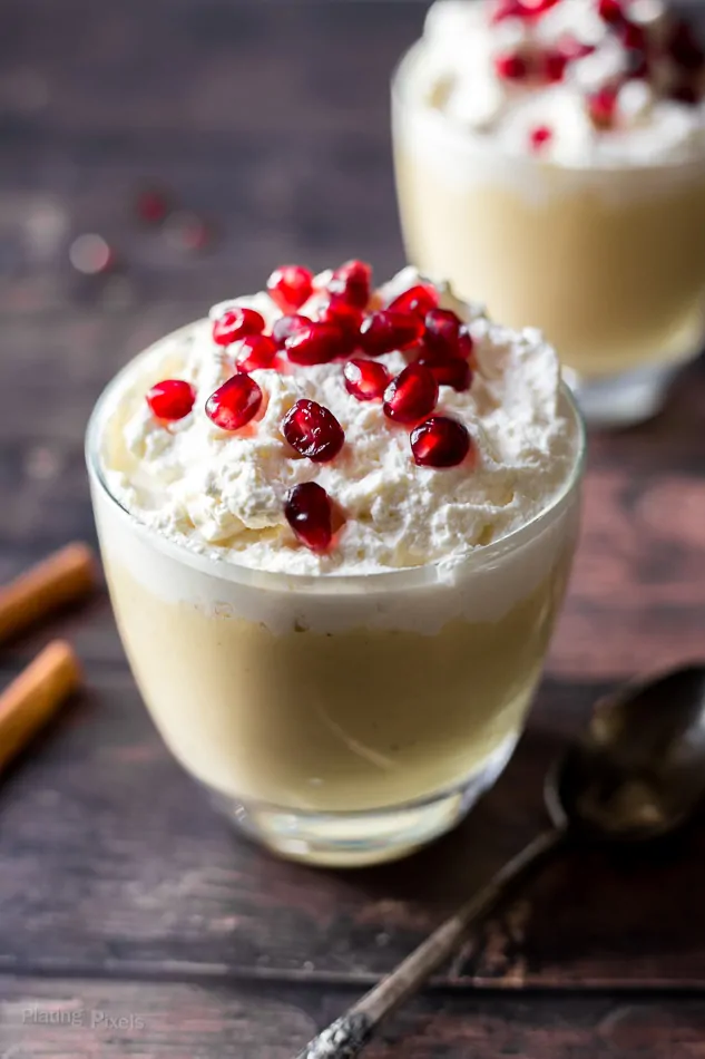 A close up of Homemade Eggnog Pudding topped with whipped cream and pomegranate sitting on a wooden surface