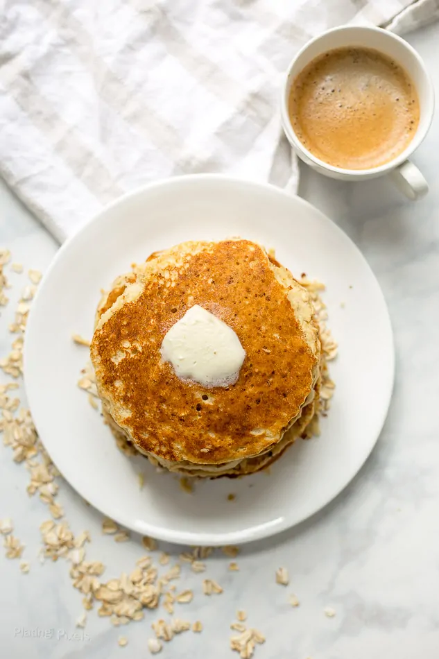An overhead shot of Banana Oatmeal Pancakes topped with butter next to a cup of coffee