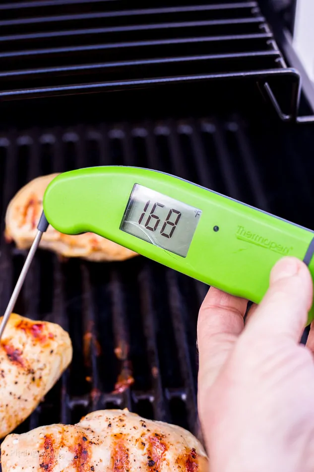 Hand holding digital thermometer in grilled chicken breast on grill to check internal temperature