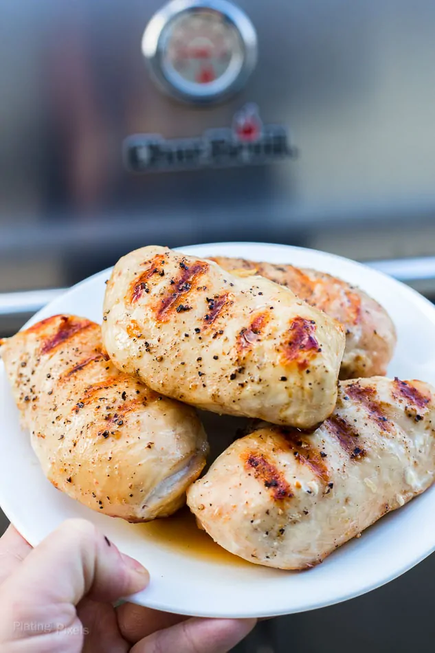 Hand holding plate of moist grilled chicken breasts next to a gas grill 
