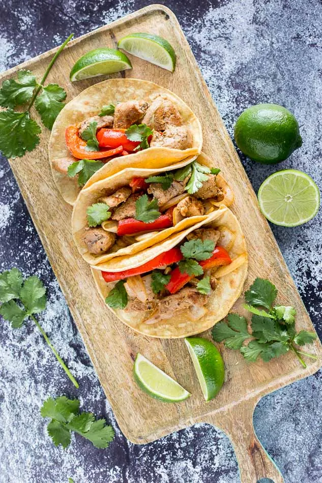 Three prepared Pork Fajita Tacos on a serving board over a gray marble backdrop surrounded by garnishes 