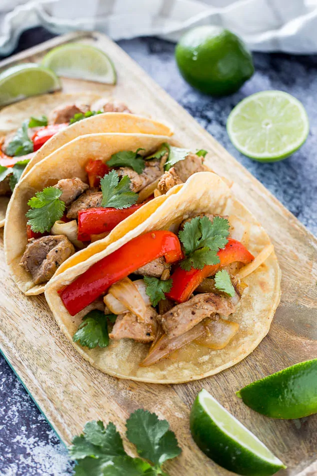 Close up of three prepared Pork Fajita Tacos on a serving board surrounded by garnishes 