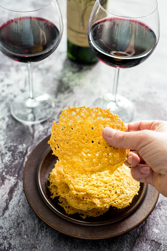 Hand holding a Parmesan Crisp with two glasses of red wine in the background