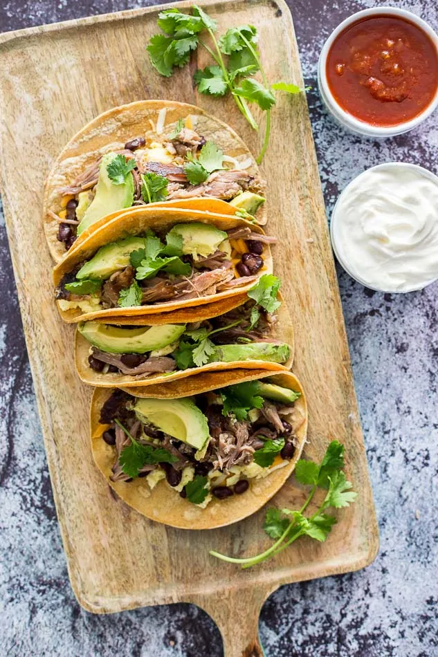 An overhead shot of Slow Cooker Carnitas Breakfast Tacos on a wooden board with salsa and sour cream