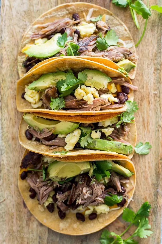 Slow Cooker Carnitas Breakfast Tacos stacked together on a wooden surface topped with avocado and cilantro