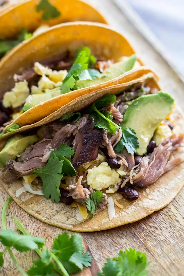 A close up of Carnitas Breakfast Tacos on a wooden surface with sliced avocado