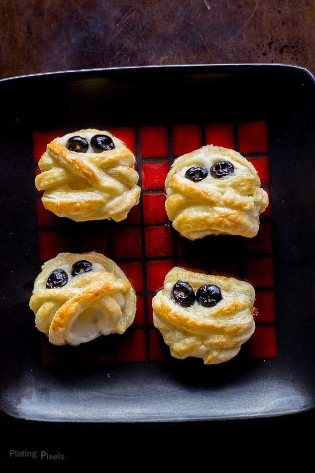 Four Halloween Puff Pastry Mummy Cheese Wheels on a red and black plate