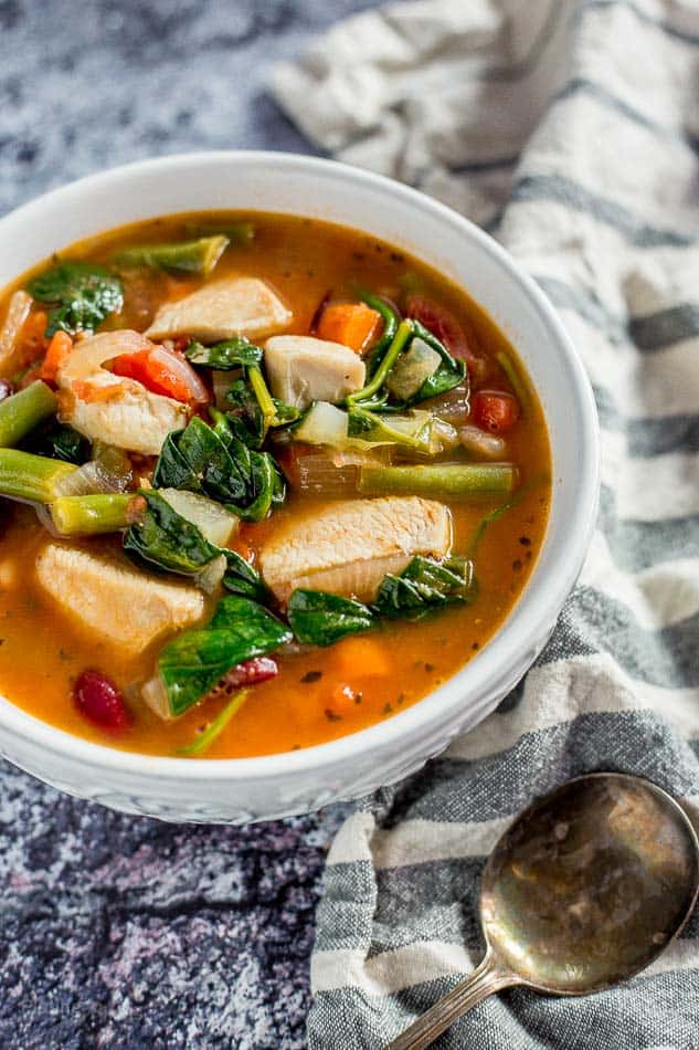 A close up of Chicken Minestrone Soup in a white bowl with green vegetables and a spoon at the side
