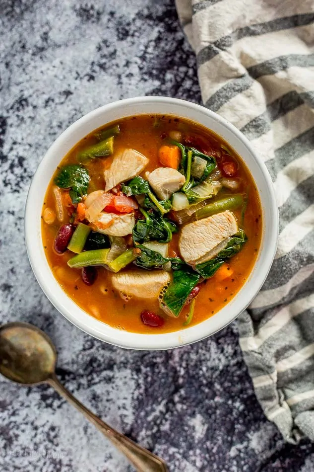 An overhead shot of Chicken Minestrone Soup in a white bowl with a spoon beside it