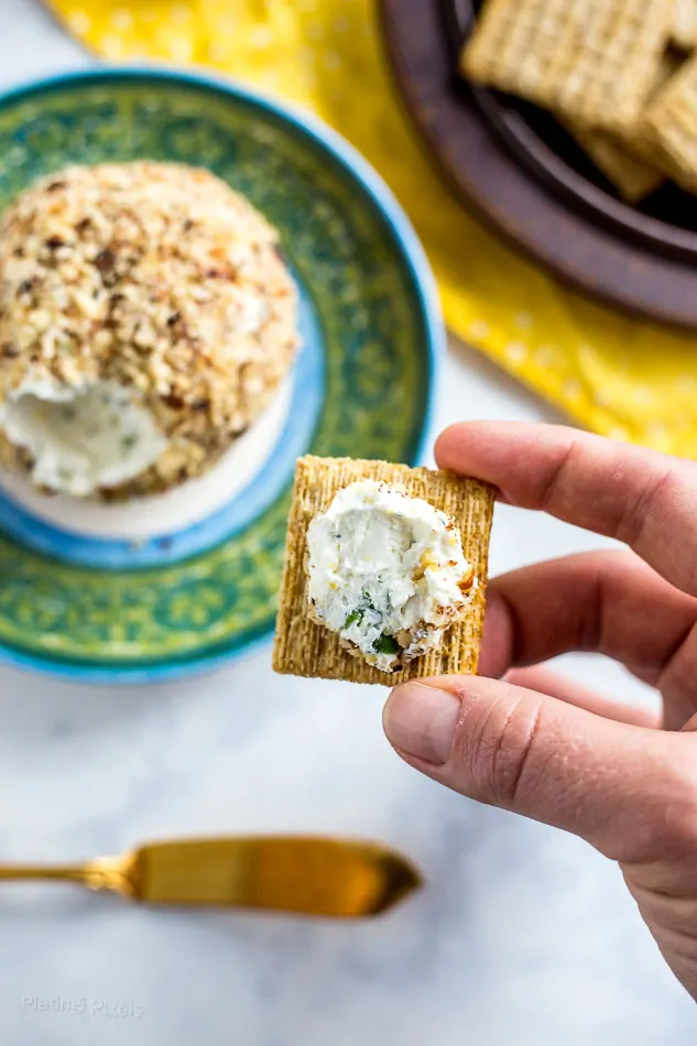 Hand holding cracker with some of Lemon Herb Goat Cheese Ball spread onto it