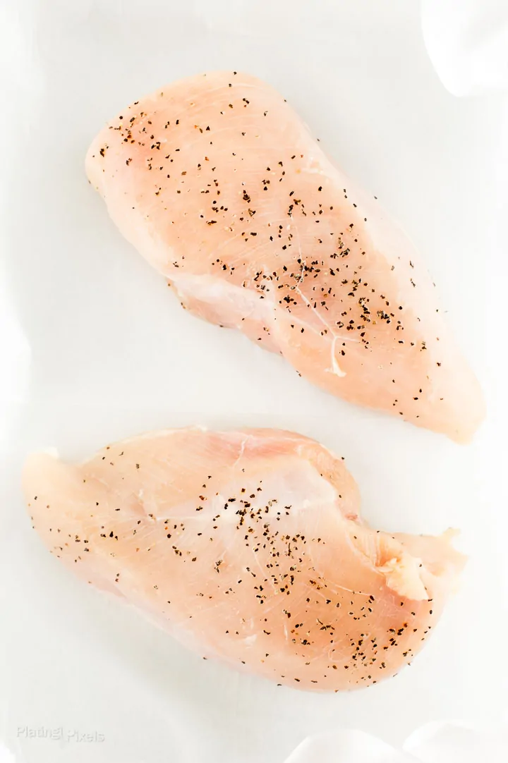 Two raw chicken breasts seasoned with salt and pepper resting at room temp