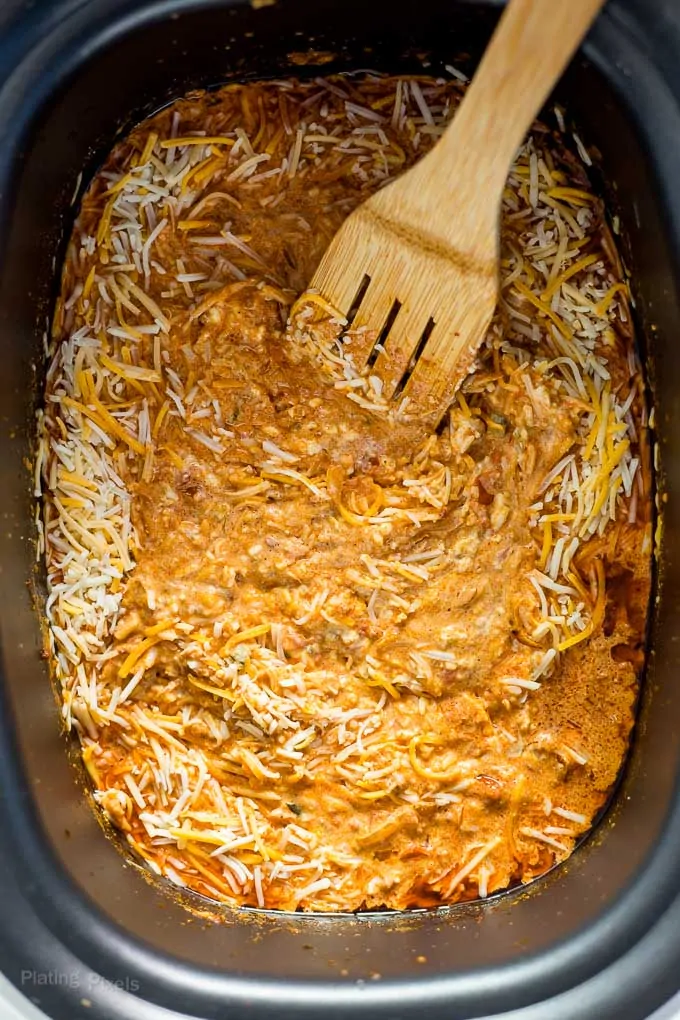 Wooden spoon stirring shredded cheese into queso dip base in a slow cooker