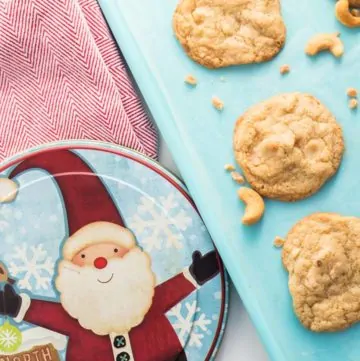 Chewy Cashew Toffee Cookies on teal cookie sheet with a santa decorated cookie tin beside it