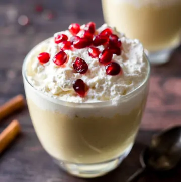A close up of Homemade Eggnog Pudding topped with whipped cream and pomegranate sitting on a wooden surface