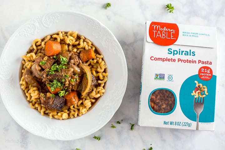 A bowl of Slow Cooker Beef Stew next to a package of Modern Tabel Lentil Spiral Pasta