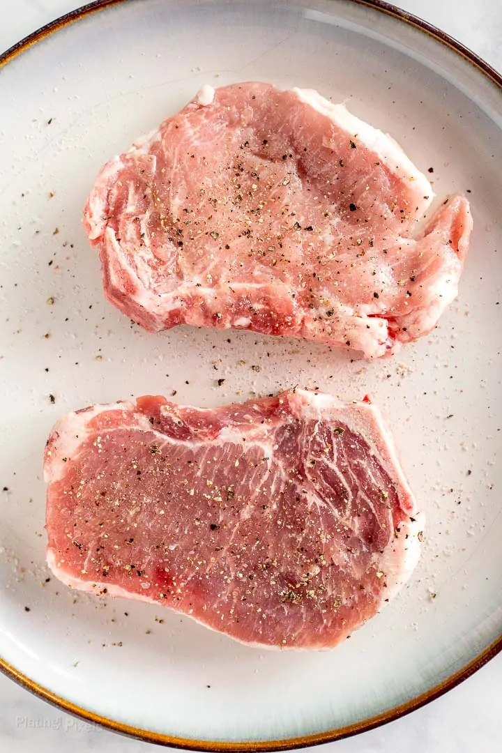 Two seasoned raw pork chops sitting on a plate at room temp