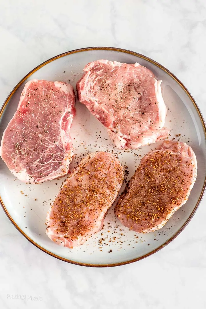 Four seasoned raw pork chops sitting on a plate at room temp