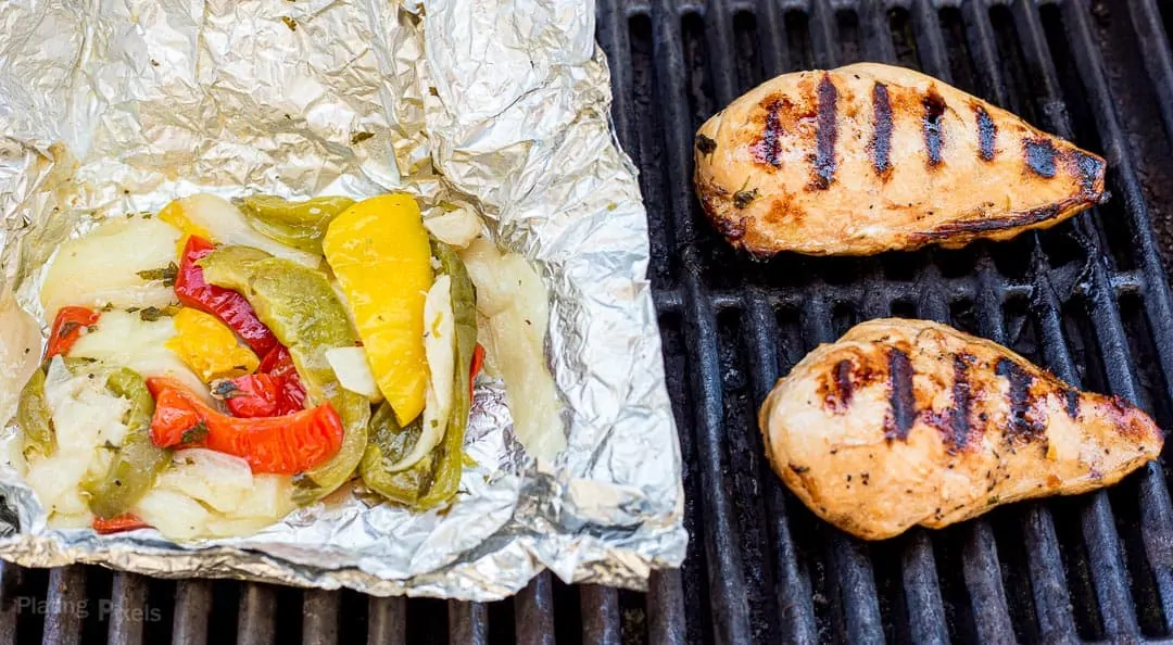 A pouch of fajita marinated vegetables cooking next to chicken breasts on a grill